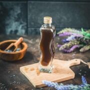 Curved bottle filled with homemade lavender extract on brown wooden board surrounded by lavender flowers.