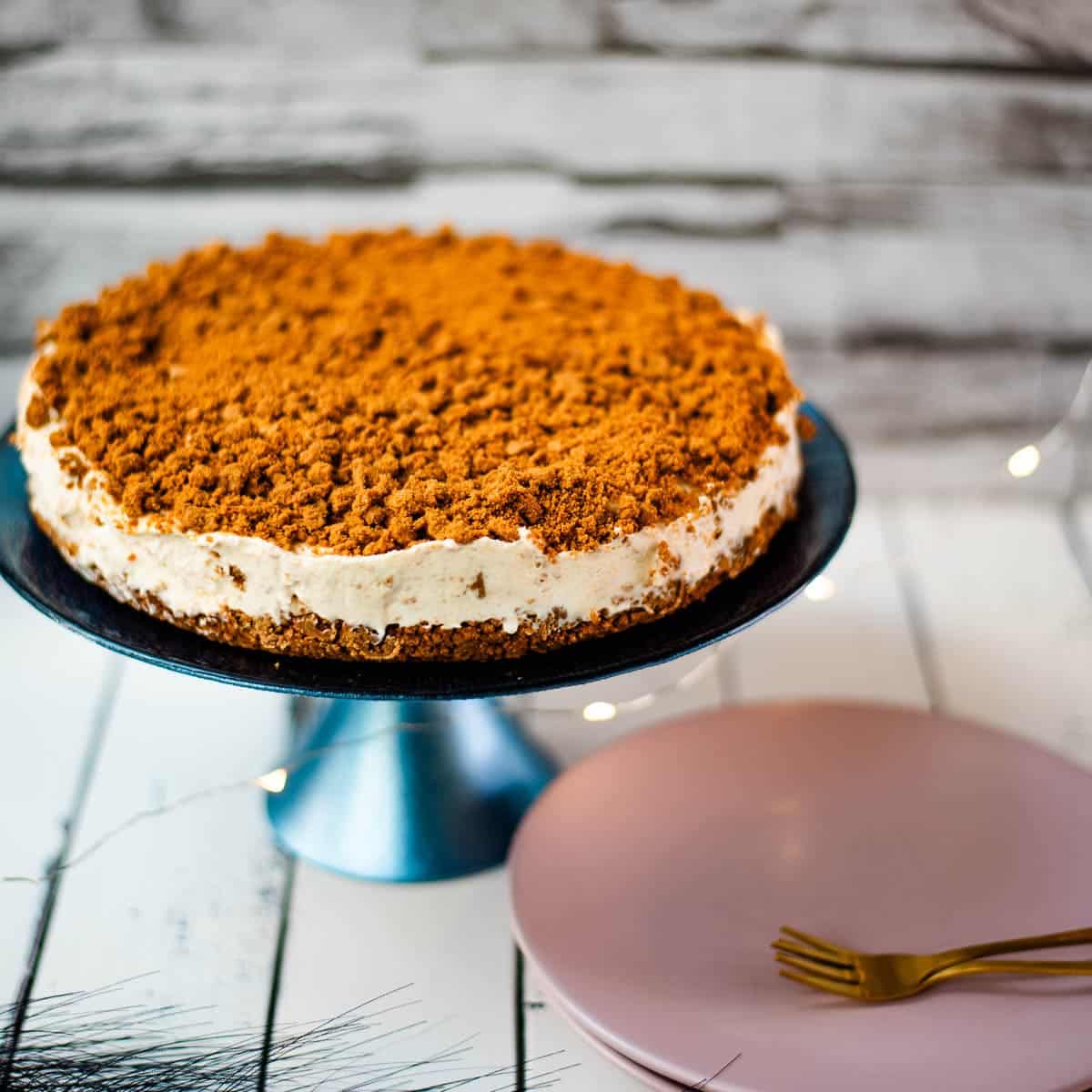 Gorgeous no bake Biscoff cheesecake on blue cake stand. Placed on white wooden ground next to two pink plates.