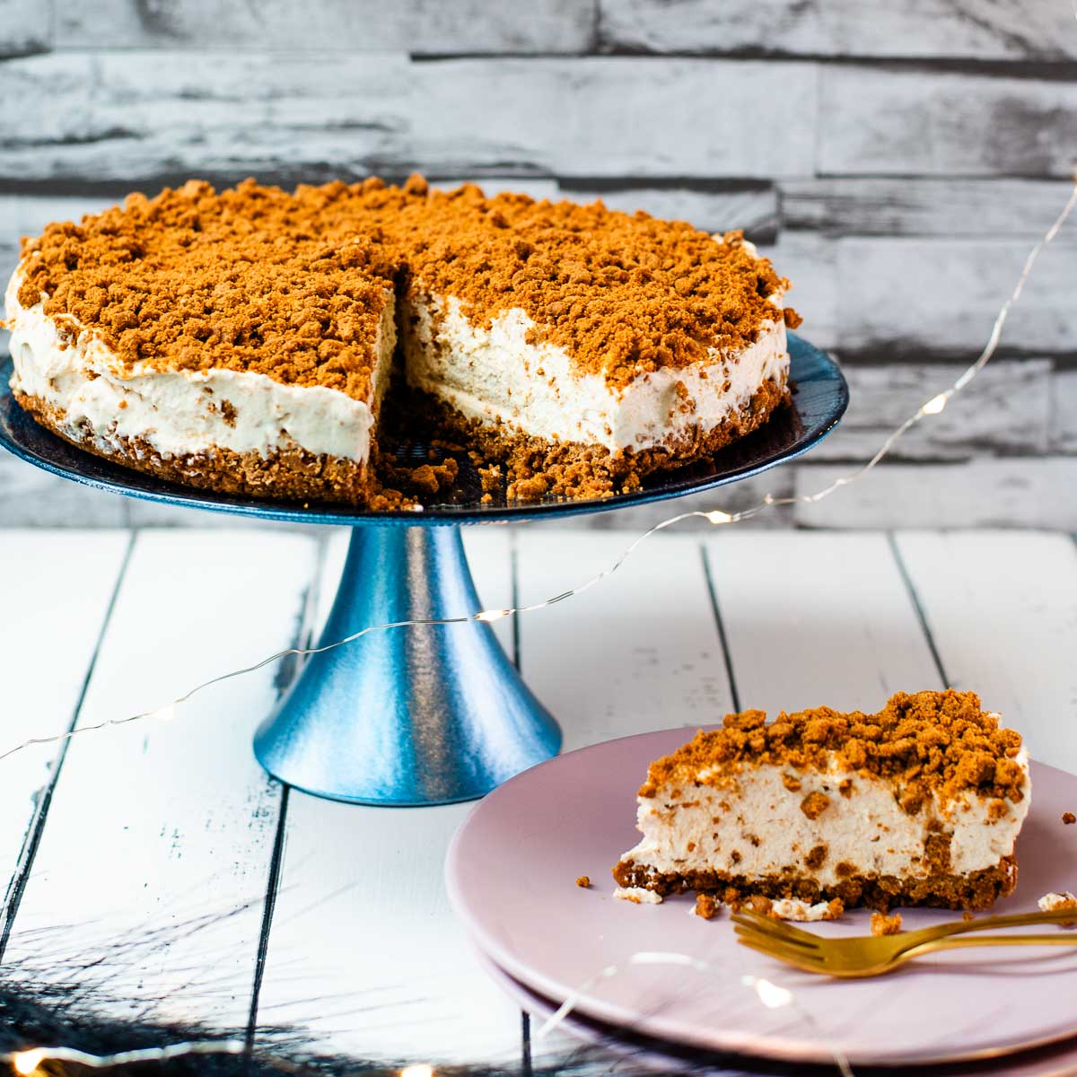 Gorgeous no bake Biscoff cheesecake on blue cake stand with a slice cut out. Placed on white wooden ground next to two pink plates.