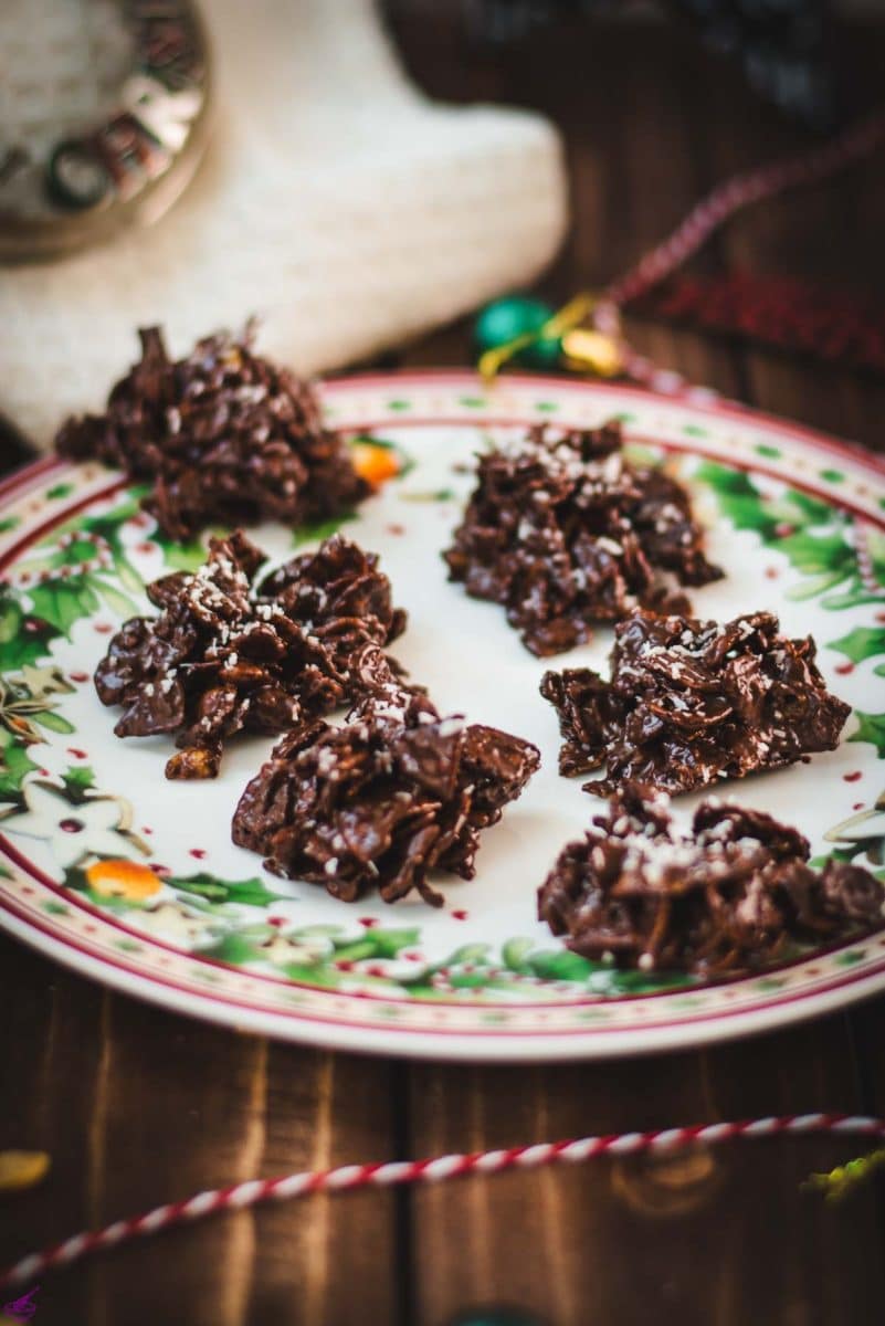 Christmas plate filled with dark chocolate clusters, on brown wooden background, next to Christmas decorations.