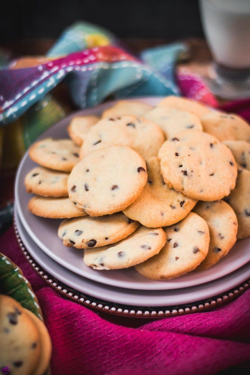 Chocolate chip sugar cookies placed on pink plates, embedded in colorful tea towels, in front of a glass of milk.