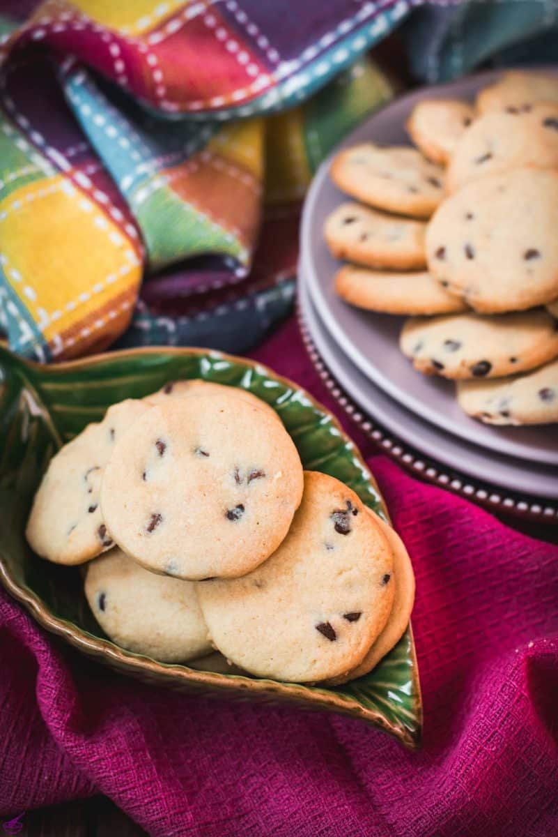 Chocolate chip sugar cookies placed on pink plates, embedded in colorful tea towels, in front of a glass of milk.