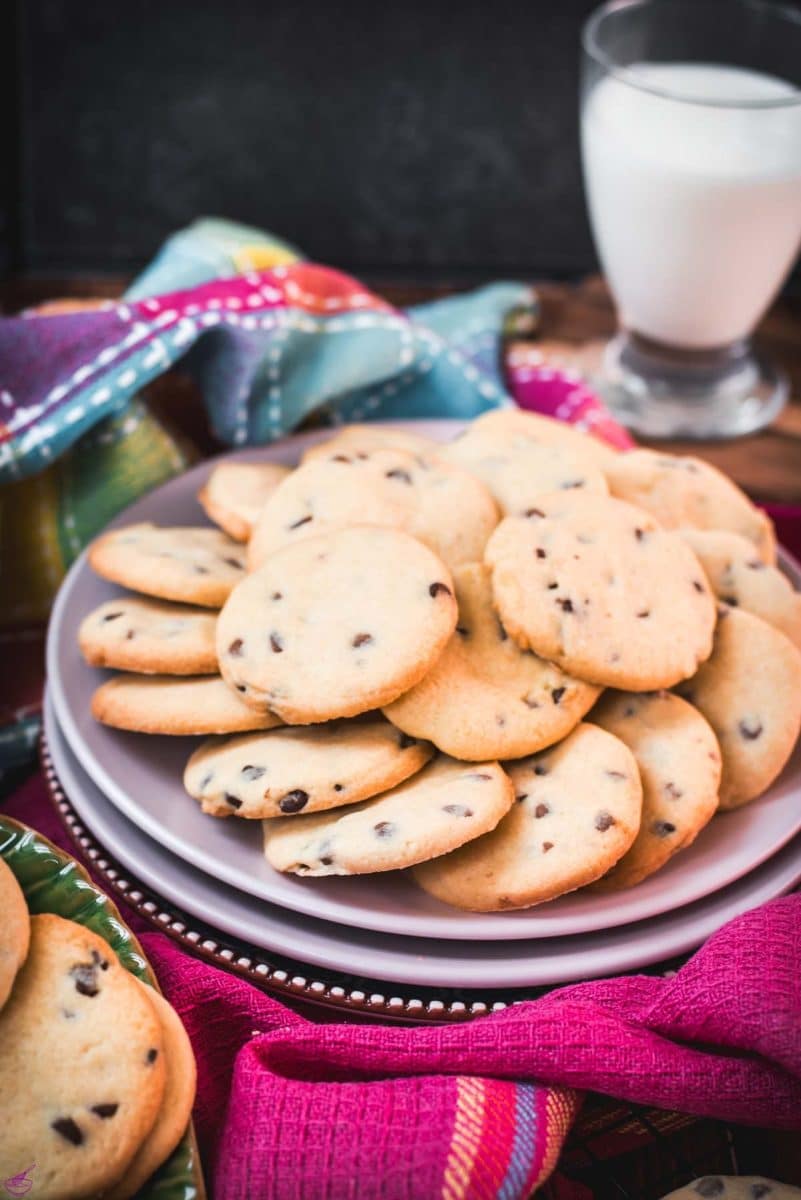 Chocolate chip sugar cookies placed on pink plates, embedded in colorful tea towels, in front of a glass of milk.