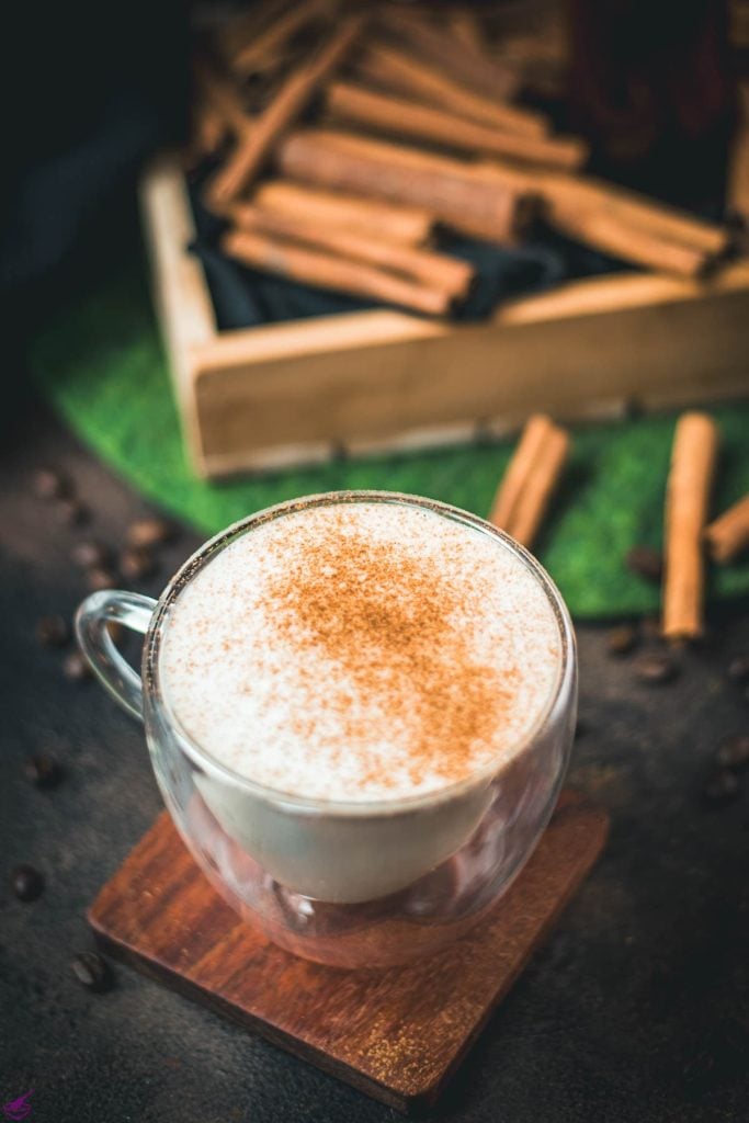 Gorgous cinnamon dolce latte, dusted with ground cinnamon. Placed on wooden coaster with cinnamon sticks in the background.