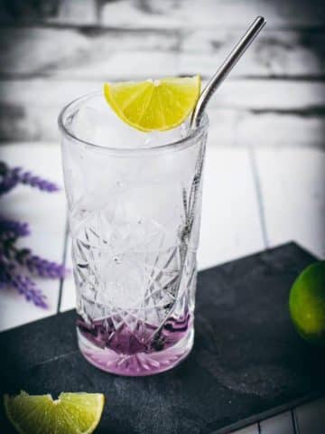 Highball glass filled with lavender gin and tonic, garnished with lime wedge and straw. Placed on black slate plate.