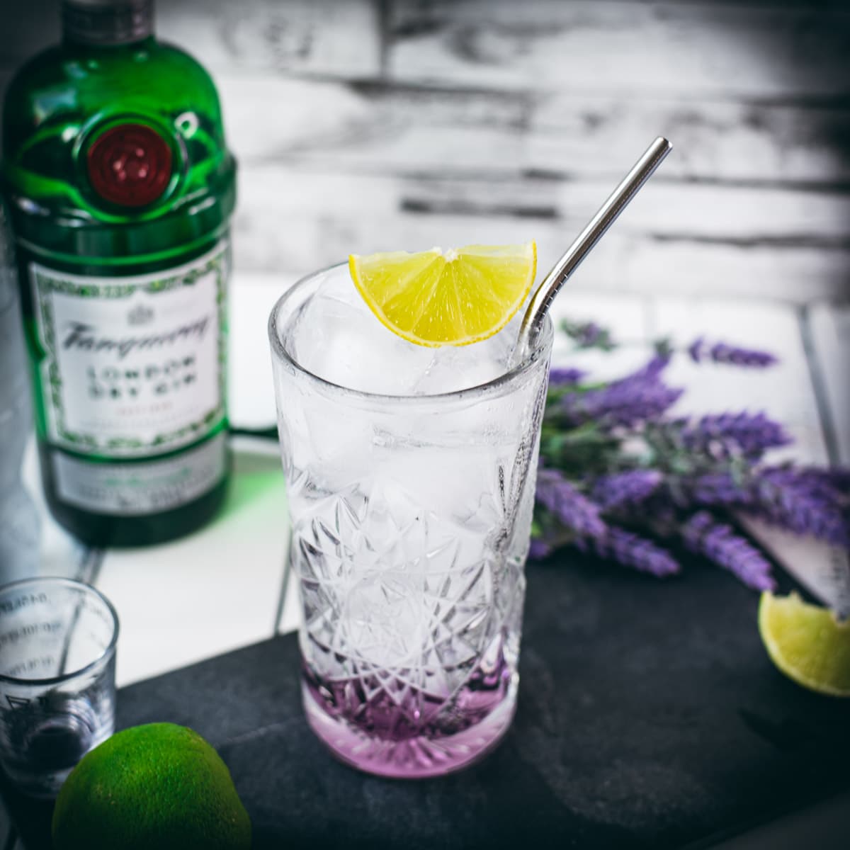 Highball glass filled with lavender gin and tonic, garnished with lime wedge and straw. Placed on black slate plate.