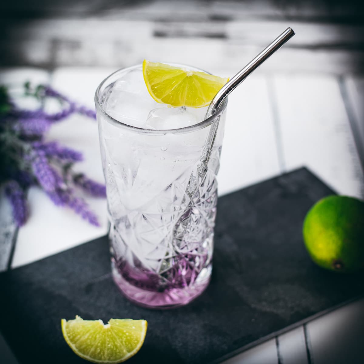 Highball glass filled with lavender gin and tonic, garnished with lime wedge and straw. Placed on black slate plate.