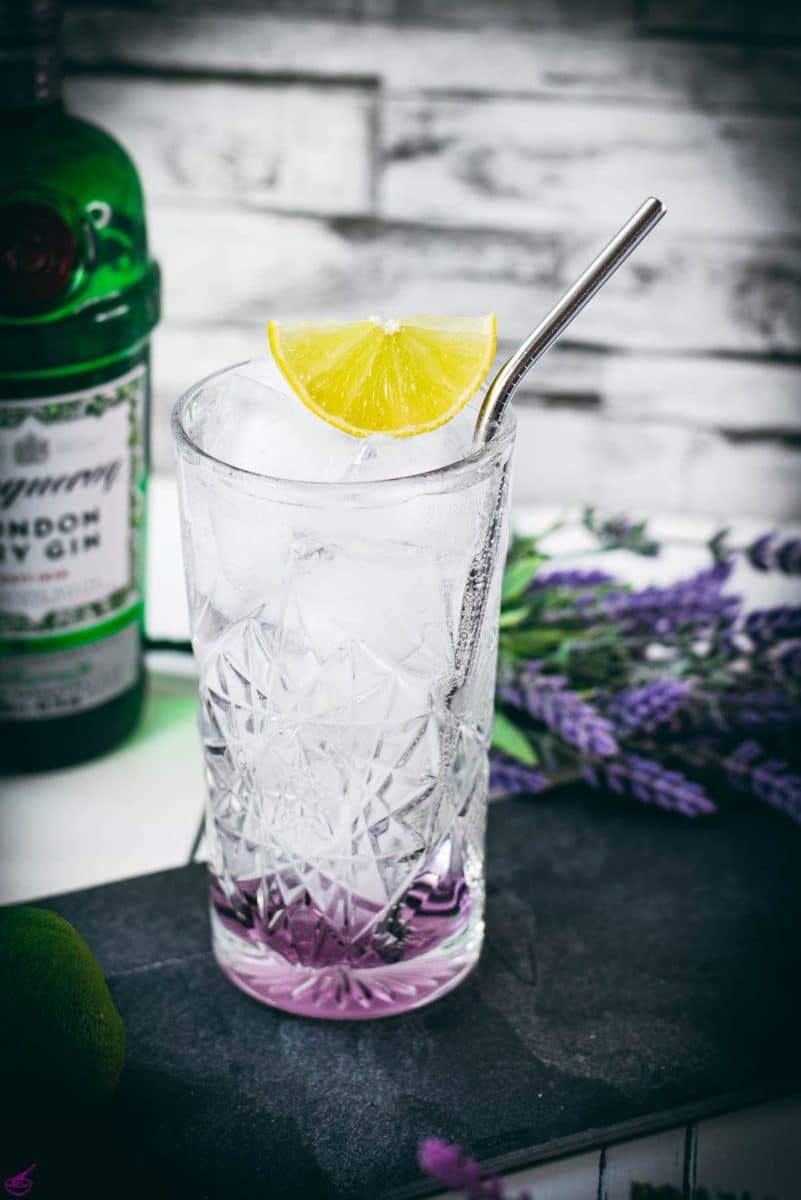 Highball glass filled with lavender gin and tonic, garnished with lime wedge and straw. Placed on black slate plate.