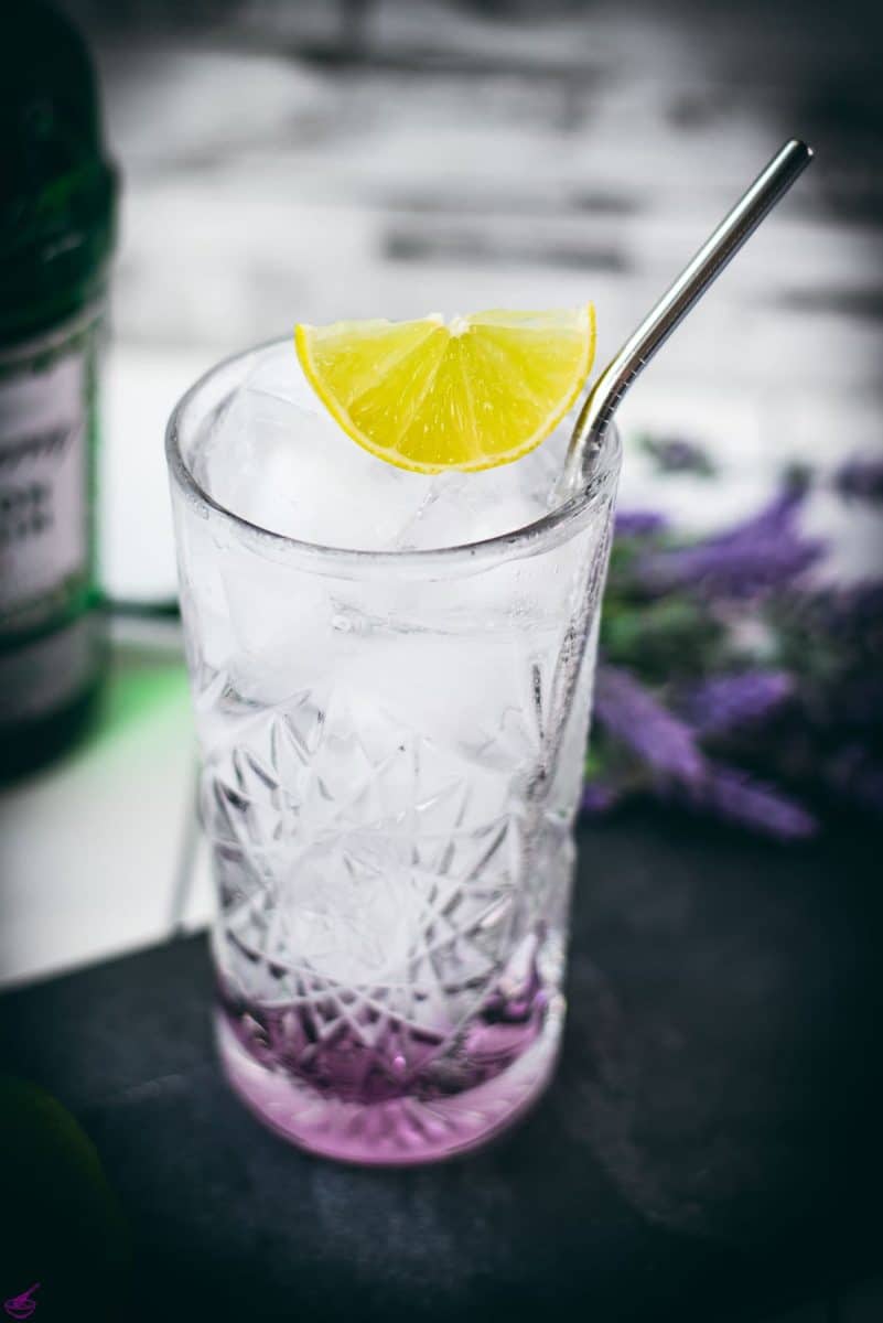 Highball glass filled with lavender gin and tonic, garnished with lime wedge and straw. Placed on black slate plate.