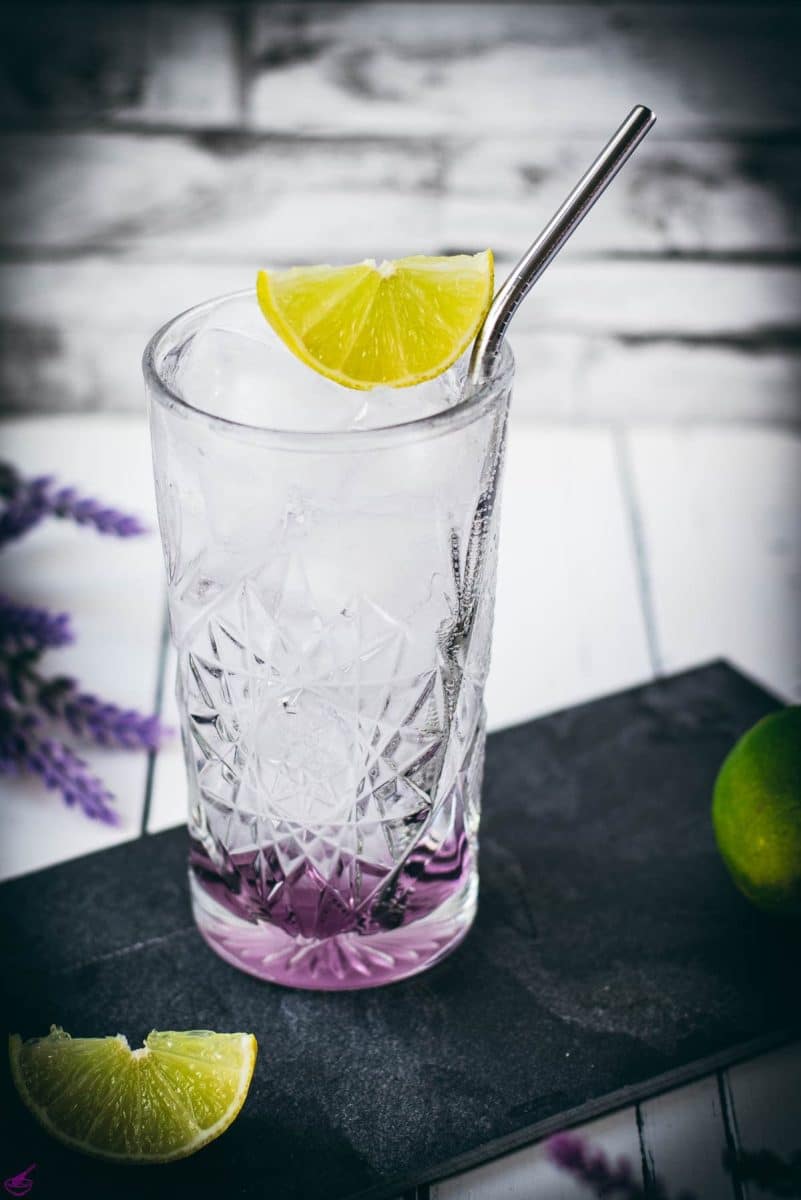 Highball glass filled with lavender gin and tonic, garnished with lime wedge and straw. Placed on black slate plate.