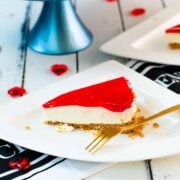 A slice of delicious Valentine's Raspberry Cheesecake placed on white plate next to a golden fork .