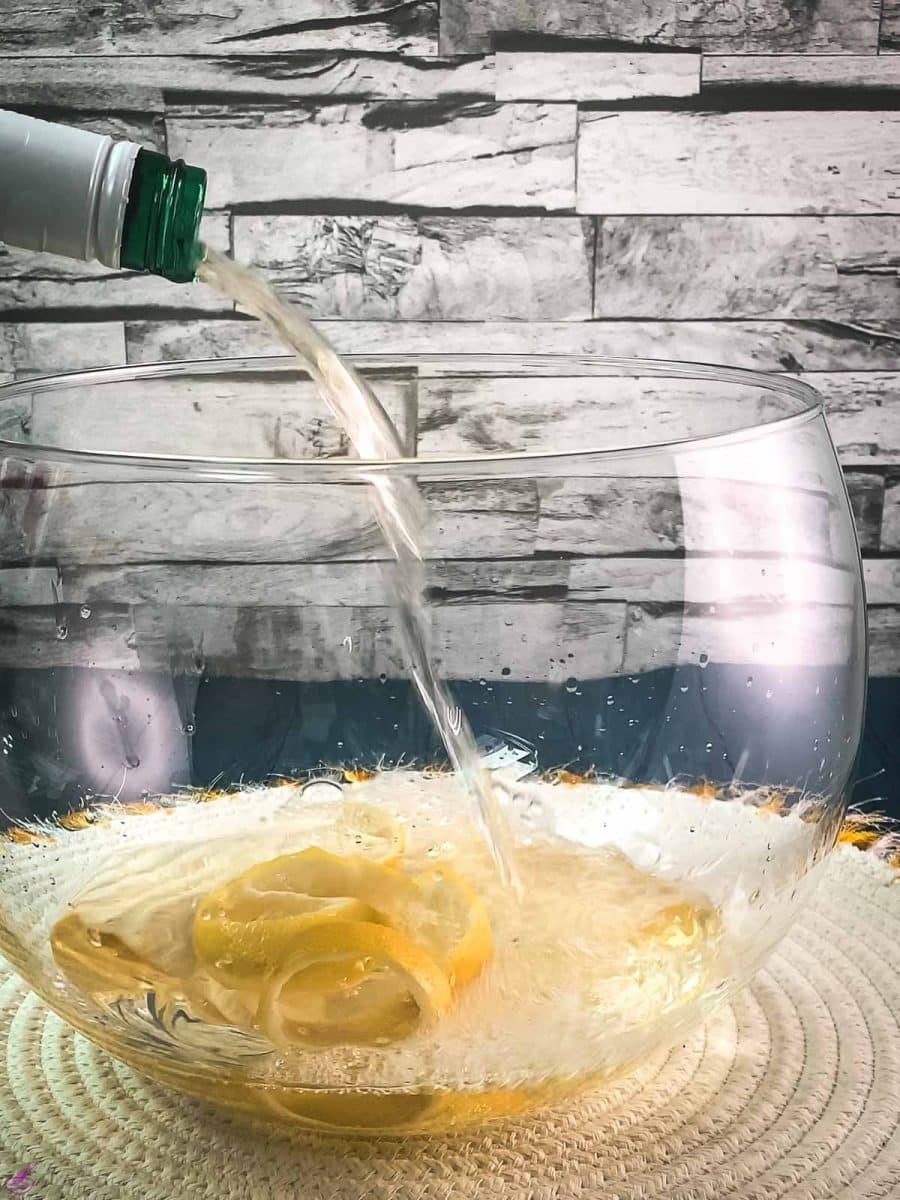 Pouring white wine into a glass punch bowl, filled with lemon zest. placed on white table mat.