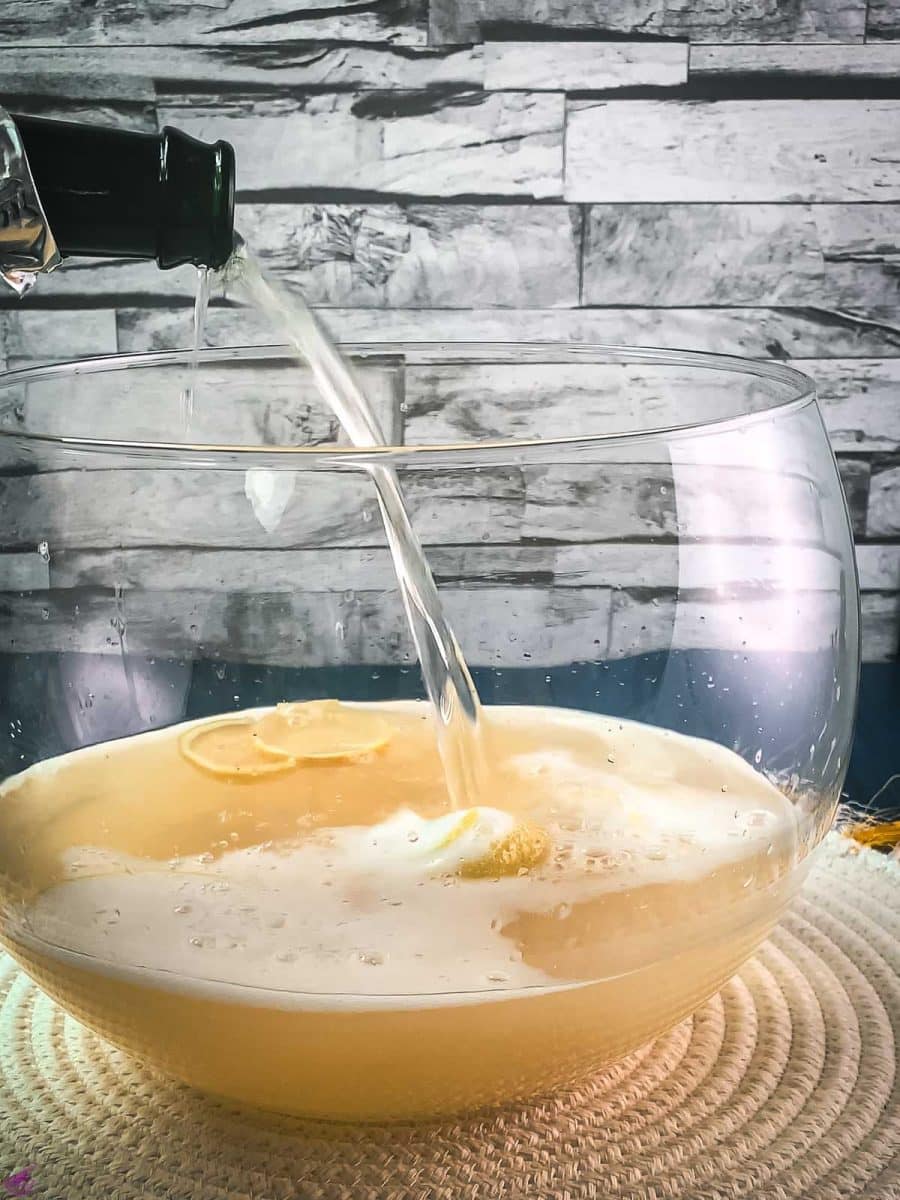 Pouring sparkling wine into a glass punch bowl, filled with white wine, lemon juice, and lemon zest. placed on white table mat.