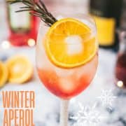 Gorgeous wine glass filled with Winter Aperol spritz, placed on snowy ground.