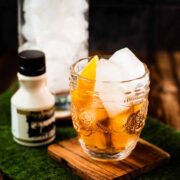 Gorgeous glass filled with maple old fashioned on wooden coaster. The drink is garnished with an orange zest and placed next to a stirring glass and a maple syrup bottle.