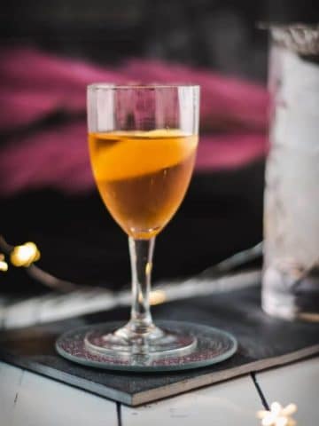 Glass filled with a delicious Martinez cocktail, garnished with a lemon zest. Placed on a black slate.