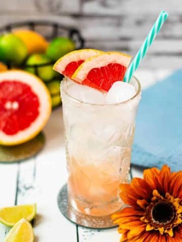 Colorful and refreshing paloma cocktail ina a highball glass, placed on mirrored coaster. Garnished with grapefruit slices and a straw.