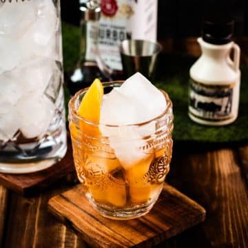 Gorgeous glass filled with maple old fashioned on wooden coaster. The drink is garnished with an orange zest and placed next to a stirring glass and a maple syrup bottle.