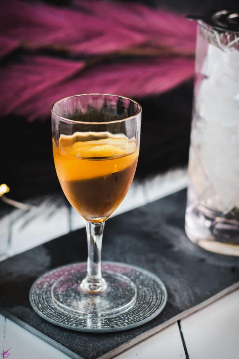 Glass filled with a delicious Martinez cocktail, garnished with a lemon zest. Placed on a black slate.