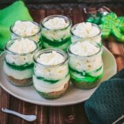 Gorgeous no bake lime cheesecakes in a jar for celebrating St. Patrick's Day! Placed on a white plate sitting on a brown wooden floor.