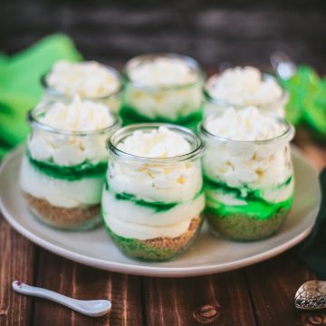 Gorgeous no bake lime cheesecakes in a jar for celebrating St. Patrick's Day! Placed on a white plate sitting on a brown wooden floor.