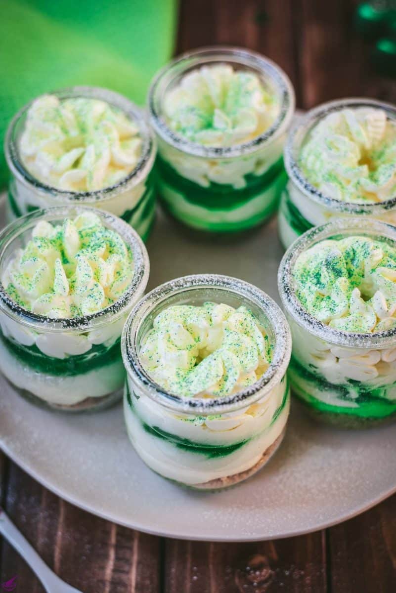 Gorgeous no bake lime cheesecakes in a jar for celebrating St. Patrick's Day! Placed on a white plate sitting on a brown wooden floor.