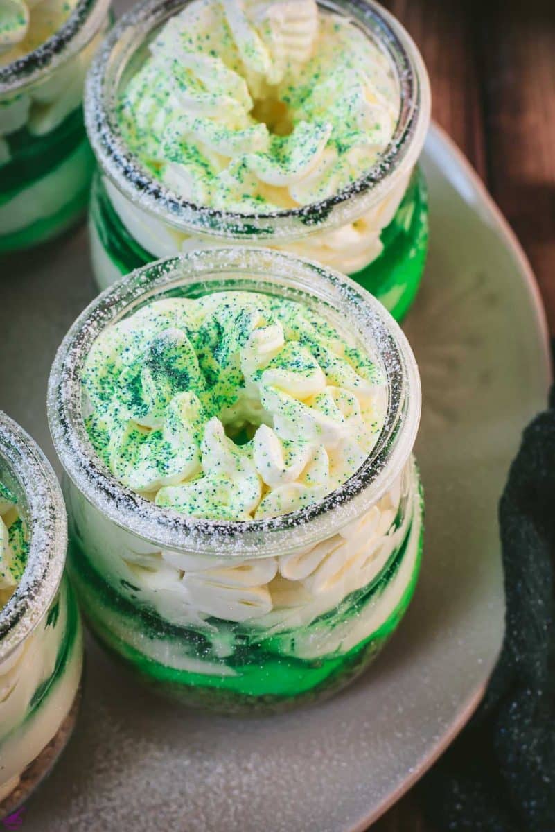 Gorgeous no bake lime cheesecakes in a jar for celebrating St. Patrick's Day! Placed on a white plate sitting on a brown wooden floor.