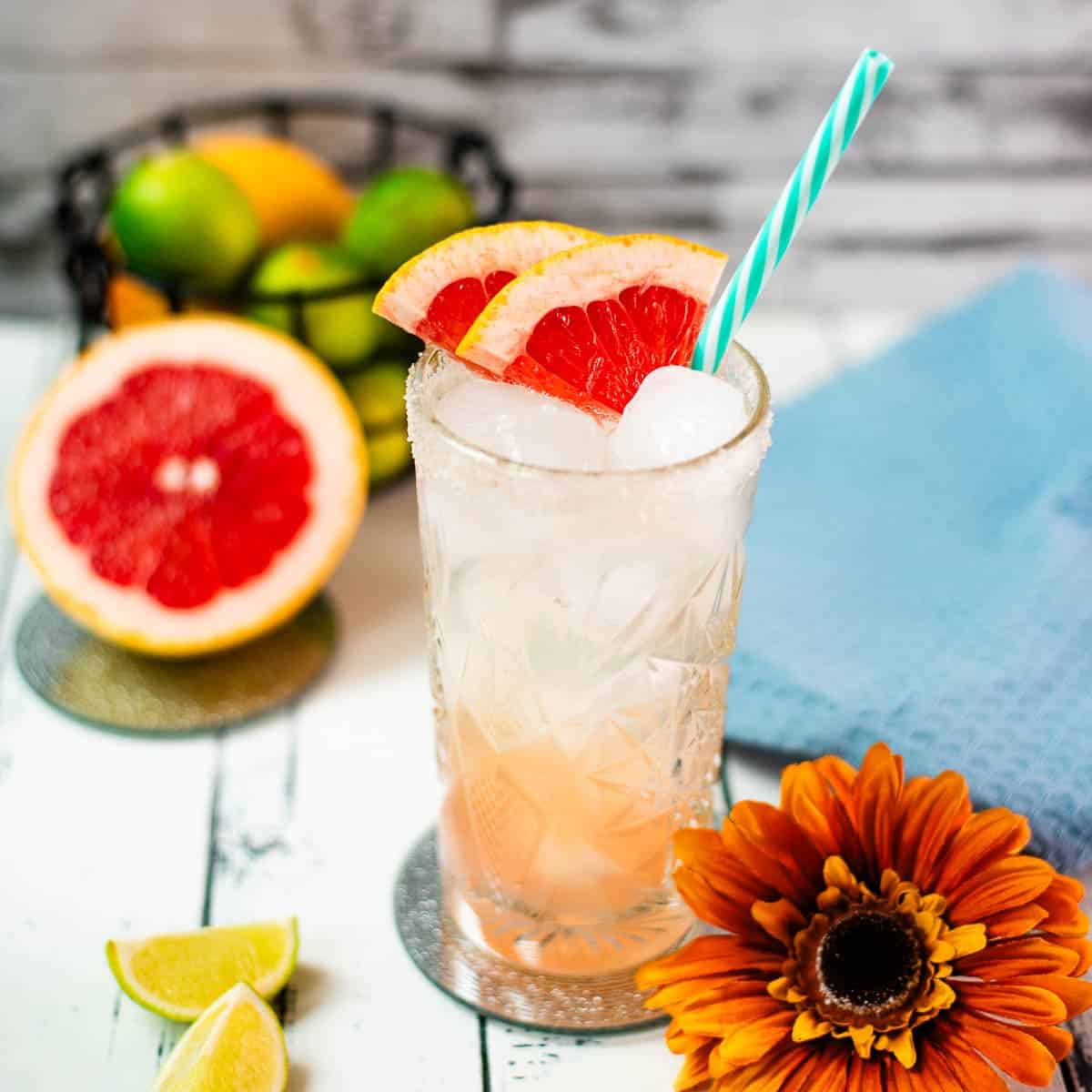 Colorful and refreshing paloma cocktail ina a highball glass, placed on mirrored coaster. Garnished with grapefruit slices and a straw.