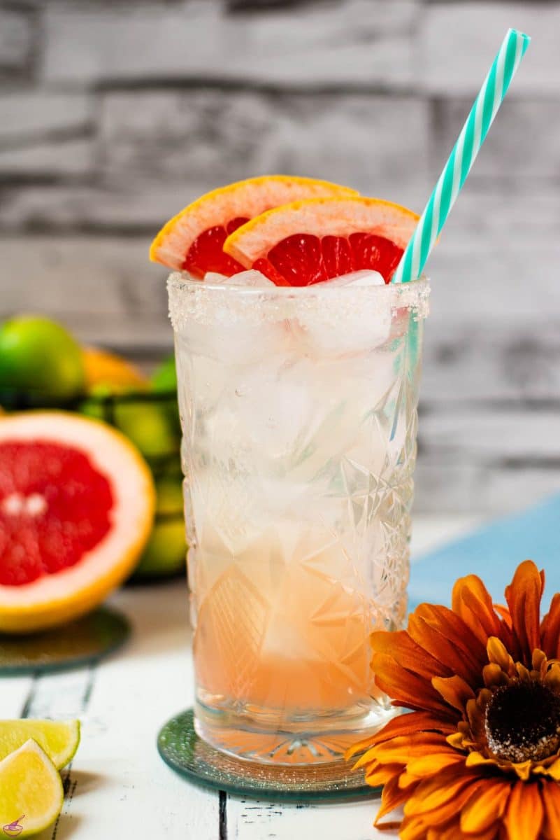 Colorful and refreshing paloma cocktail ina a highball glass, placed on mirrored coaster. Garnished with grapefruit slices and a straw.