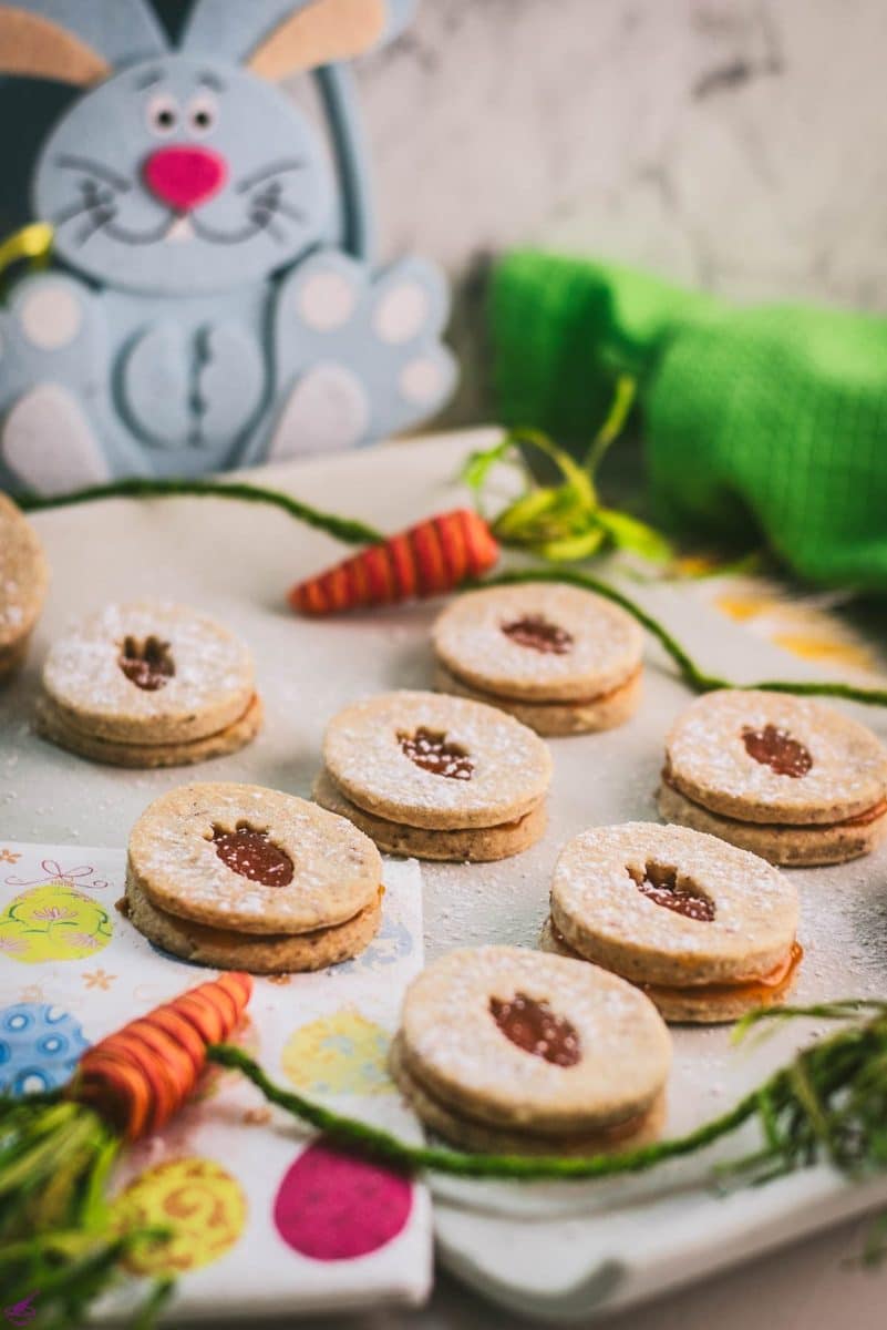 Gorgeous vegan Easter cut out cookies with apricot jam, placed on a white stone plate. A blue plush bunny smiles in the background, looking at those yummy treats.