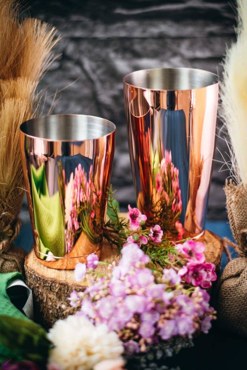 Boston shaker in rosé color, placed on wooden slice next to flowers.