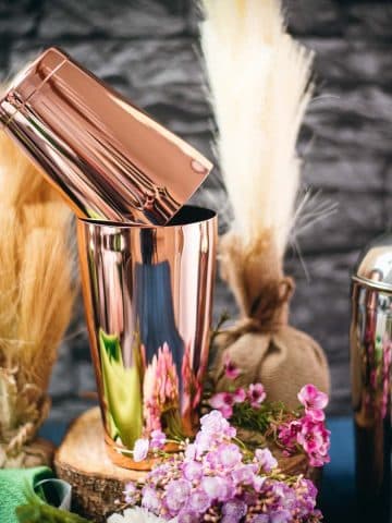 Boston shaker in rosé color, placed on wooden slice next to flowers.