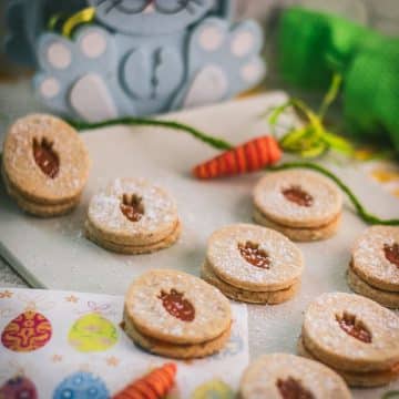 Gorgeous vegan Easter cut out cookies with apricot jam, placed on a white stone plate. A blue plush bunny smiles in the background, looking at those yummy treats.