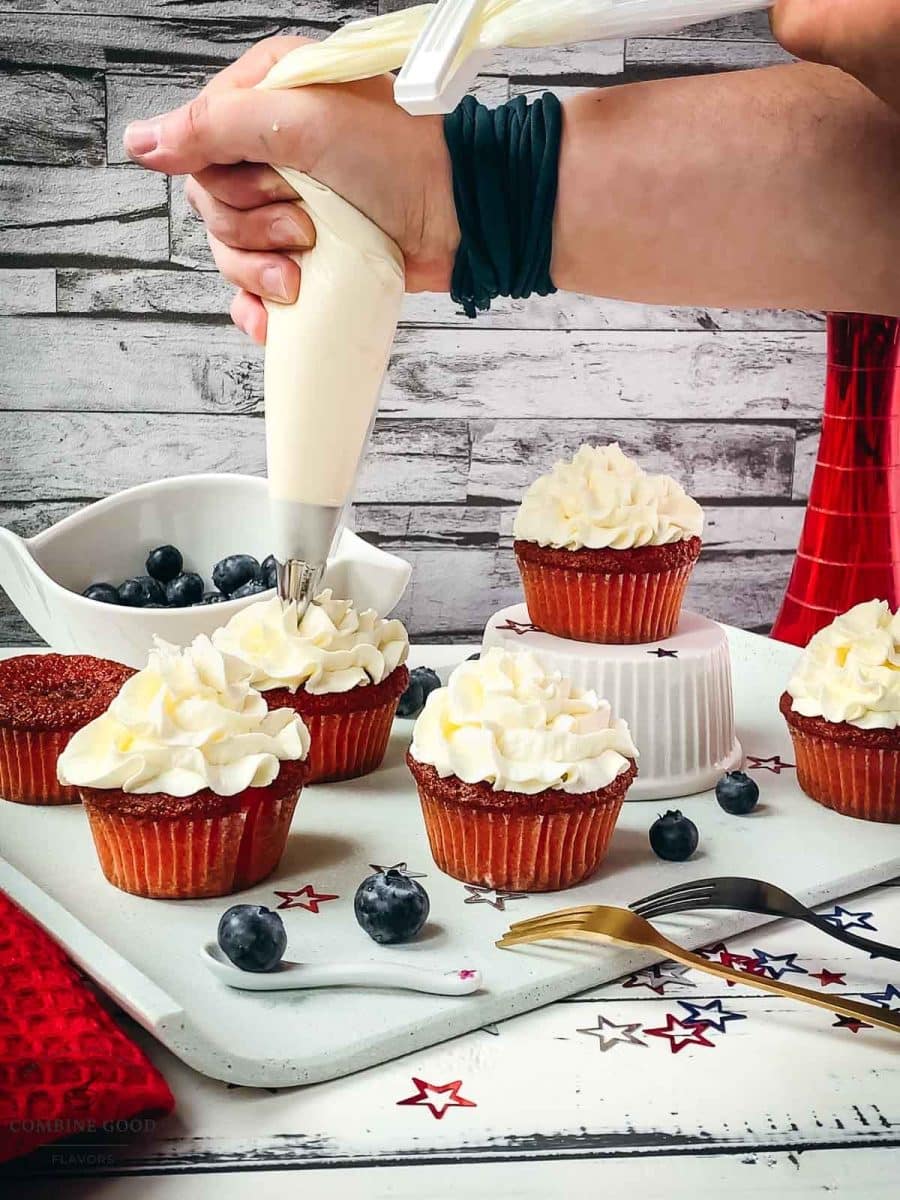 Hand piping white crème fraîche cream on top of the red muffin sponge, using a piping bag with a star tip.