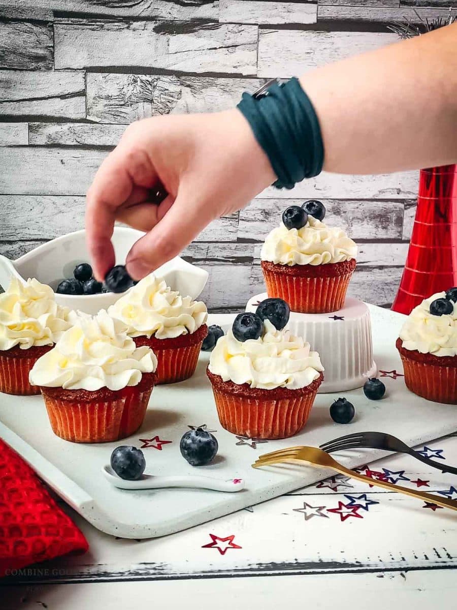 Hand placing one blueberry on top of the white cupcake topping.