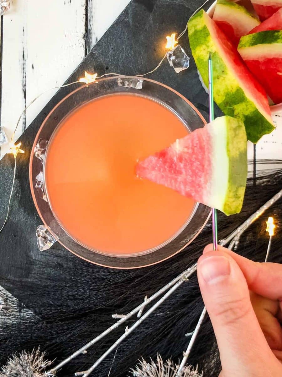 Place a watermelon slice into the drink using a cocktail pike.