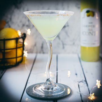 Gorgeous Limoncello vodka martini with orange sugar rim, placed on a mirrored coaster on a white wooden board.