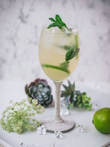 Stunning wine glass filled with Hugo spritz cocktail, placed on a white plate and garnished with mint leaves and lime slices.