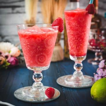 Two gorgeous glasses filled with a delicious frozen raspberry margarita cocktail, placed on glass coaster.