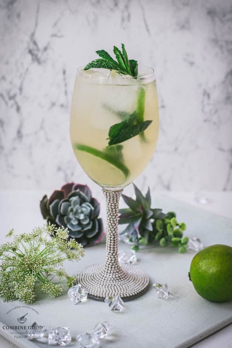 Stunning whine glass filled with Hugo spritz cocktail, placed on a white plate and garnished with mint leaves and lime slices.