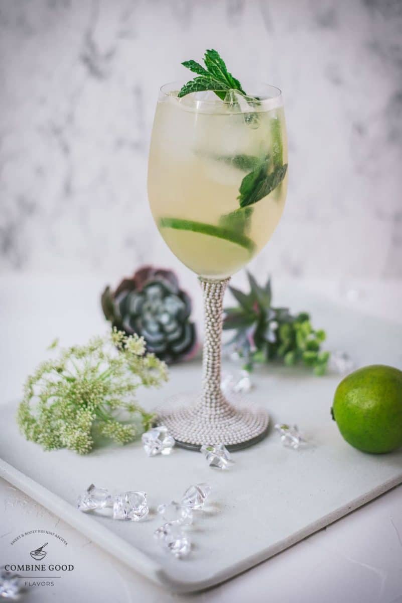 Stunning whine glass filled with Hugo spritz cocktail, placed on a white plate and garnished with mint leaves and lime slices.