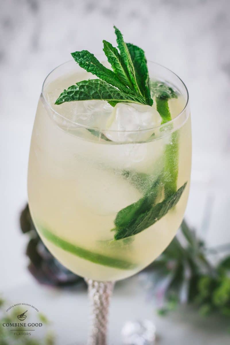 Stunning whine glass filled with Hugo spritz cocktail, placed on a white plate and garnished with mint leaves and lime slices.