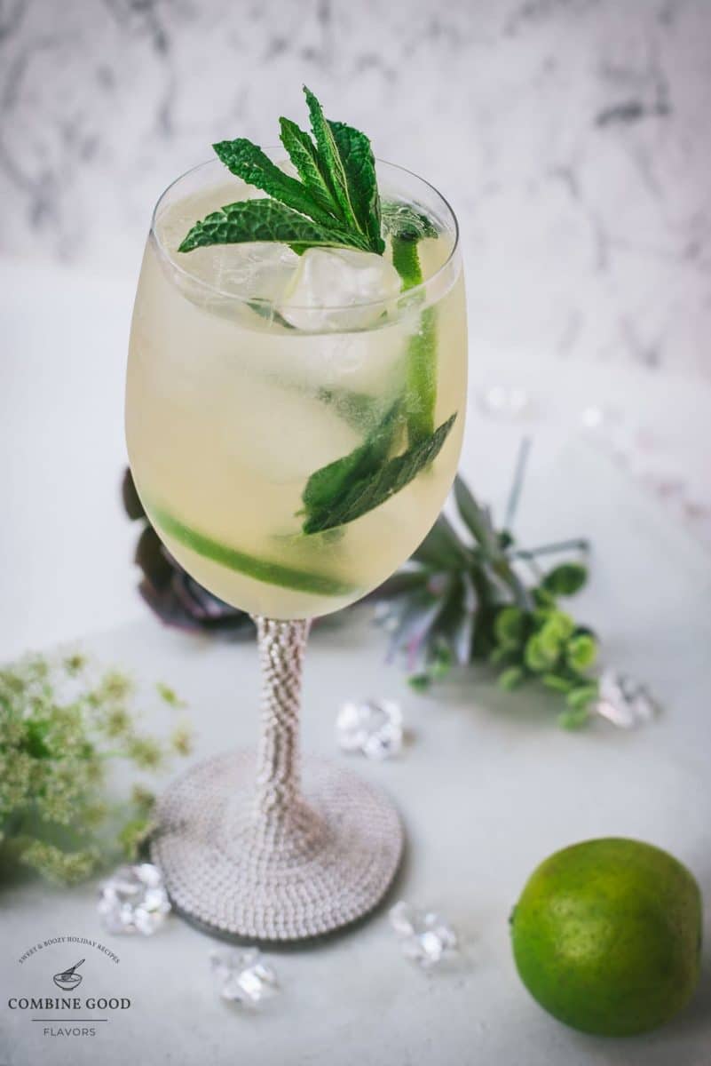 Stunning whine glass filled with Hugo spritz cocktail, placed on a white plate and garnished with mint leaves and lime slices.