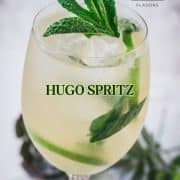 Stunning wine glass filled with Hugo spritz cocktail, placed on a white plate and garnished with mint leaves and lime slices.