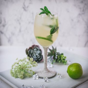 Stunning wine glass filled with Hugo spritz cocktail, placed on a white plate and garnished with mint leaves and lime slices.