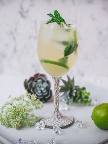 Stunning wine glass filled with Hugo spritz cocktail, placed on a white plate and garnished with mint leaves and lime slices.