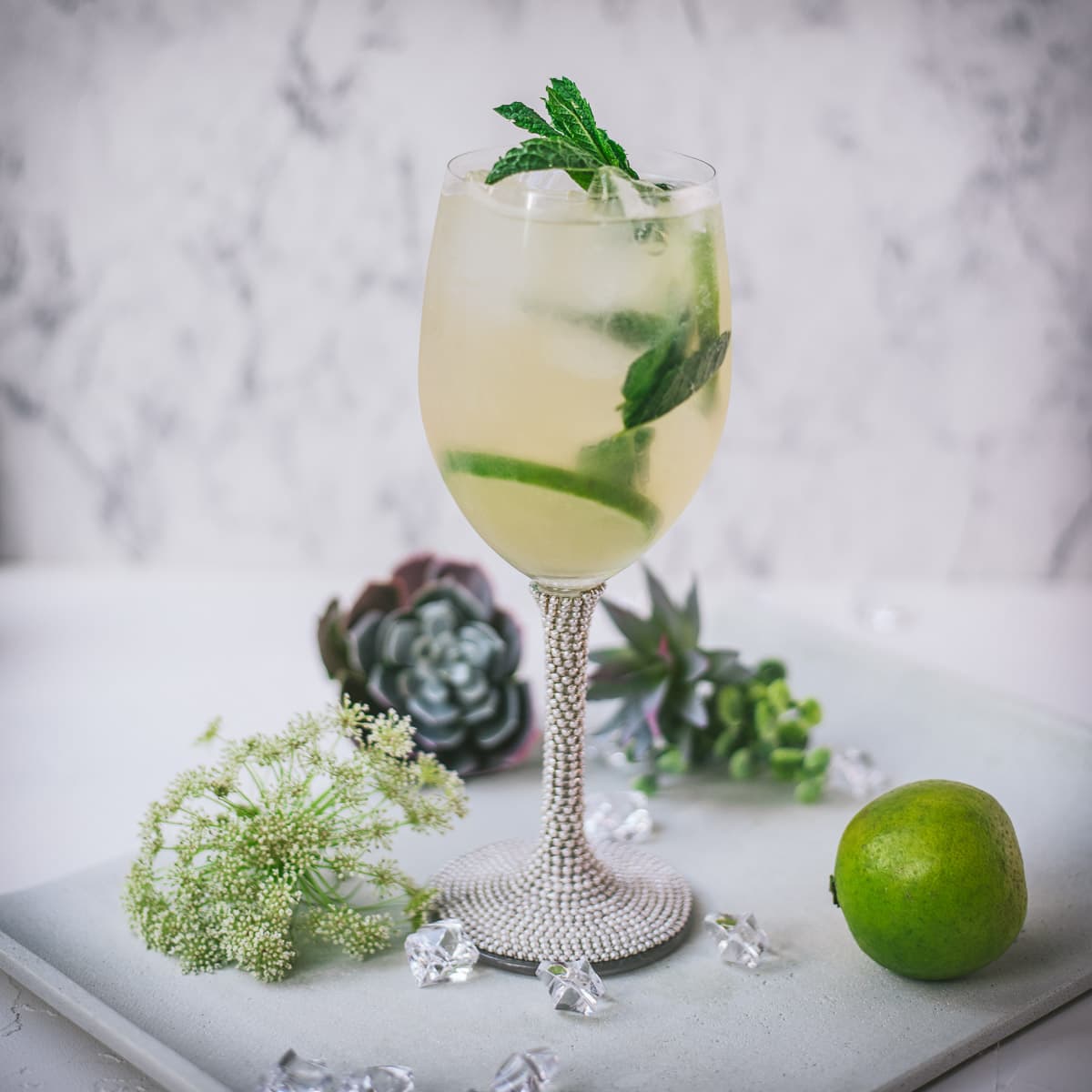 Stunning wine glass filled with Hugo spritz cocktail, placed on a white plate and garnished with mint leaves and lime slices.