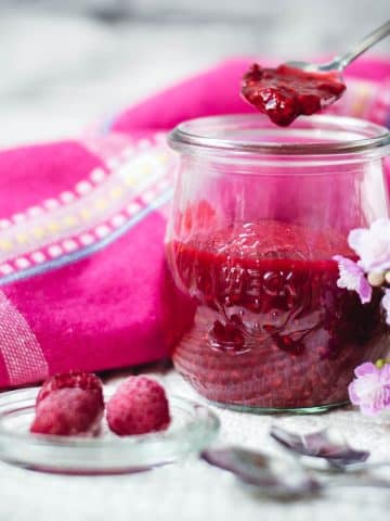 Vibrant raspberry cheesecake topping in a preserving jar, placed on white structured ground.