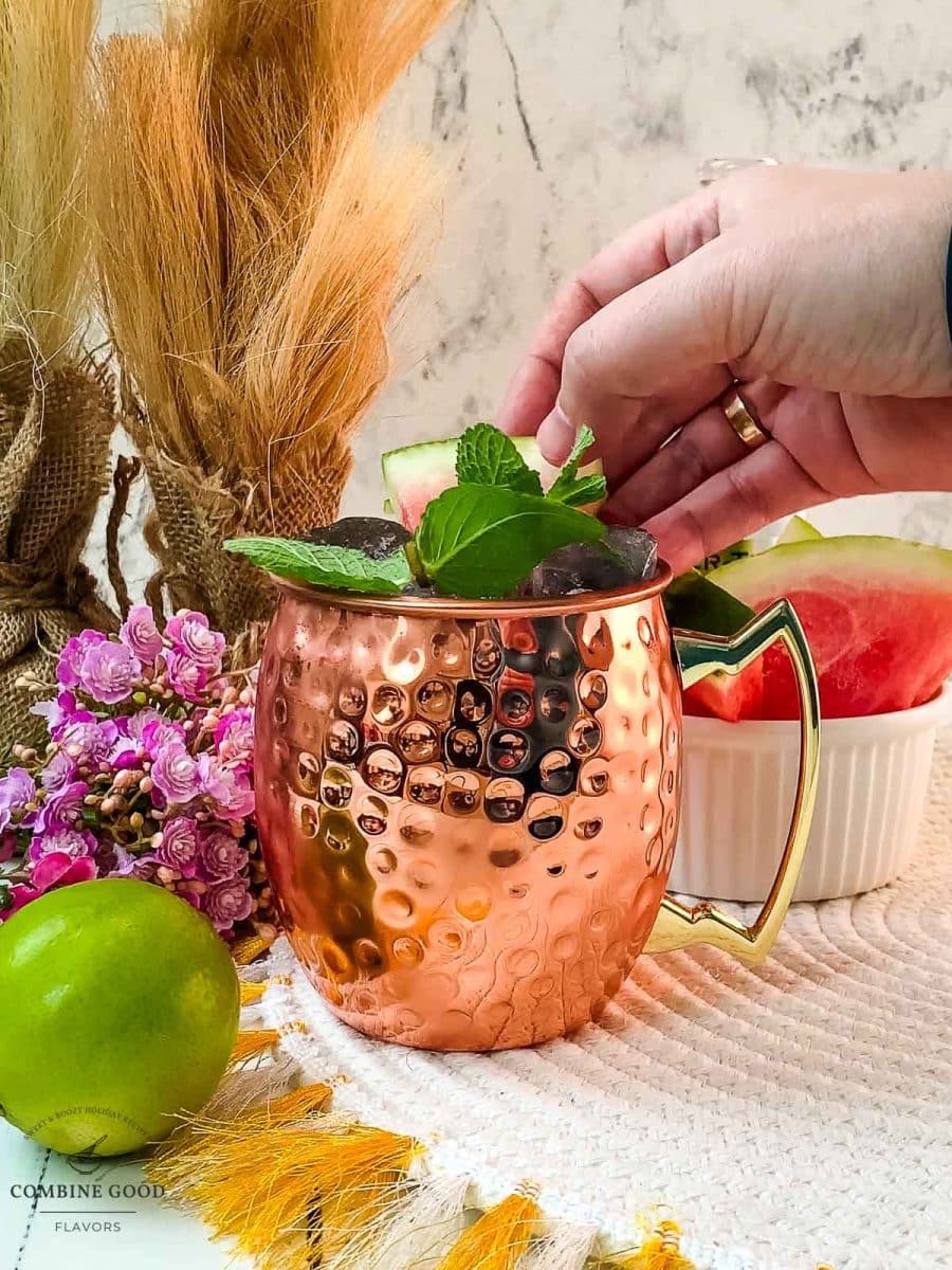 Hand placing watermelon slice into copper mug as a garnish.