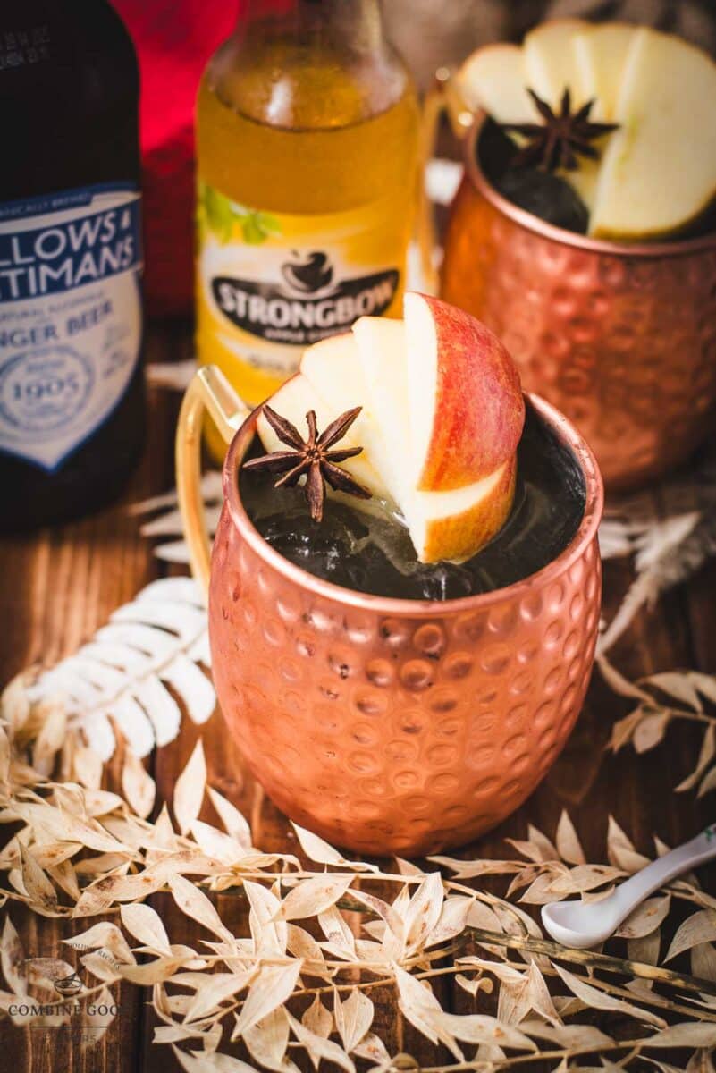 Copper mug, filled with delicious apple cider Moscow mule, garnished with fresh apple slices and a star anise. Placed on wooden board, next to a red cloth.